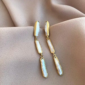 May include: A pair of long drop earrings featuring three vertical, irregular, white iridescent pearl-like segments encased in textured gold-tone metal frames. The earrings consist of a series of linked rectangular pieces that hang in a straight line.