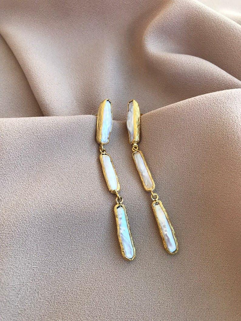 May include: A pair of long drop earrings featuring three vertical, irregular, white iridescent pearl-like segments encased in textured gold-tone metal frames. The earrings consist of a series of linked rectangular pieces that hang in a straight line.