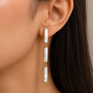 May include: A long drop earring featuring three rectangular, iridescent white mother-of-pearl segments encased in textured gold-tone metal frames. The segments are linked vertically, creating a slender, dangling design that hangs from the earlobe.
