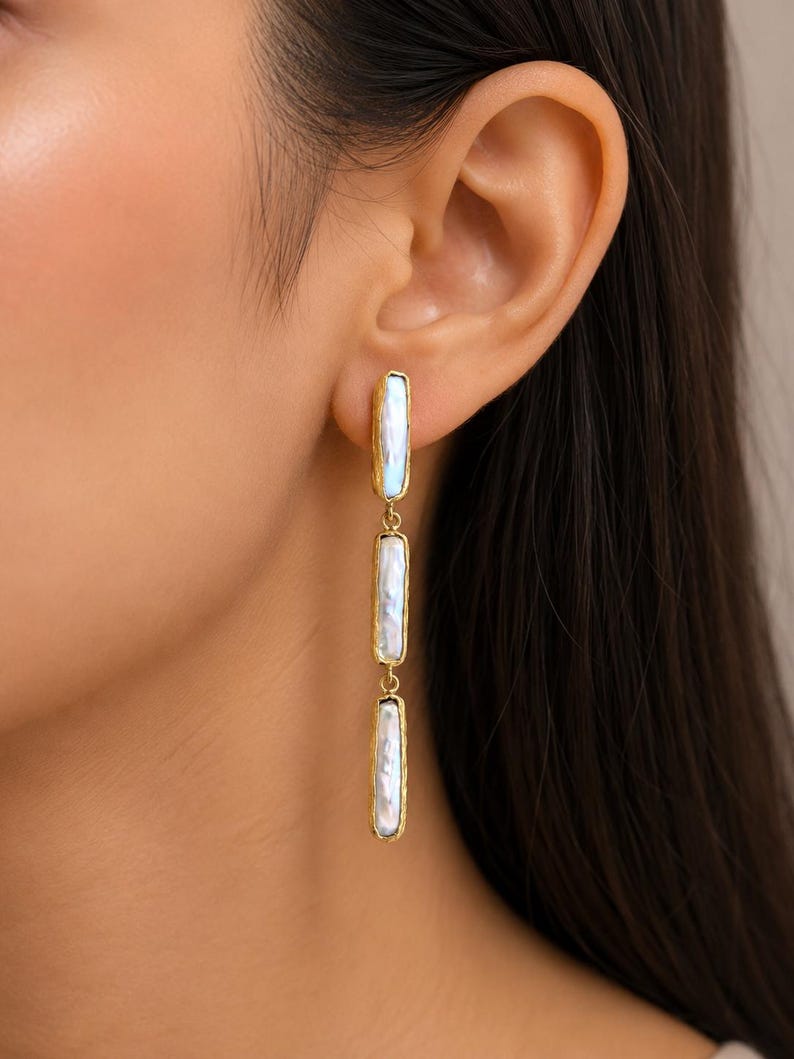 May include: A long drop earring featuring three rectangular, iridescent white mother-of-pearl segments encased in textured gold-tone metal frames. The segments are linked vertically, creating a slender, dangling design that hangs from the earlobe.