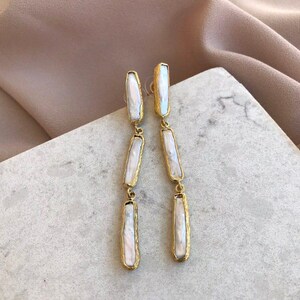 May include: A pair of long drop earrings featuring three vertical, rectangular-shaped white iridescent pearls set in textured gold-toned metal frames. Each earring consists of three linked segments that hang in a straight line from a post back.