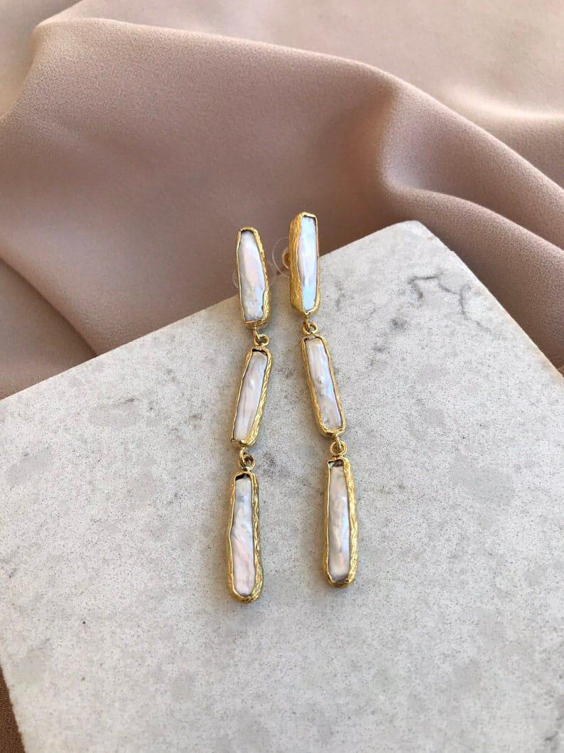 May include: A pair of long drop earrings featuring three vertical, rectangular-shaped white iridescent pearls set in textured gold-toned metal frames. Each earring consists of three linked segments that hang in a straight line from a post back.