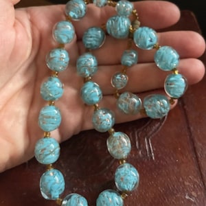 May include: A turquoise glass bead necklace with gold flecks. The beads are round and have a textured surface. The necklace is strung on a brown cord.