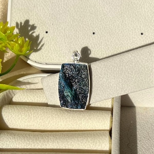 Könnte beinhalten: Ein rechteckiger Anhänger mit einem dunklen, strukturierten Stein in tiefblauen und schwarzen Farbtönen, eingefasst in einen schlichten silberfarbenen Metallrahmen mit einer kleinen runden Öse an der Oberseite zur Befestigung, präsentiert in einer Schmuckschatulle.