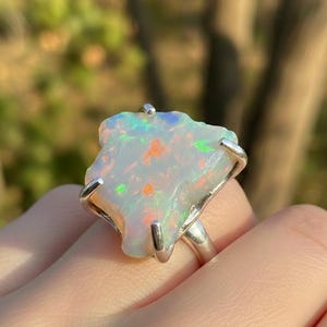 May include: A silver ring featuring a large, irregular, raw opal gemstone with a white base and vibrant flashes of orange and green color play. The stone is secured in a four-prong setting, showcasing its natural, unpolished, and jagged edges.