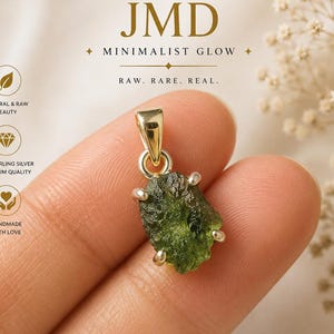 May include: A raw green moldavite stone pendant set in a polished silver-toned metal bail with four prongs. The text JMD MINIMALIST GLOW RAW. RARE. REAL. RAW MOLDAVITE PENDANT WEAR NATURE. WEAR CONFIDENCE. is visible along with icons for natural beauty.