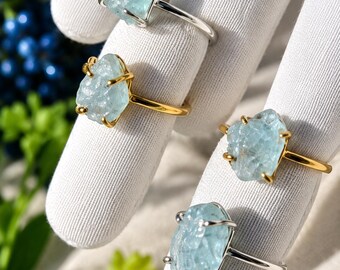 May include: Four rings featuring raw, light blue, translucent stones set in metal bands. Two rings have silver-toned bands and two rings have gold-toned bands. Each stone is held in place by four small metal prongs on the ring setting.