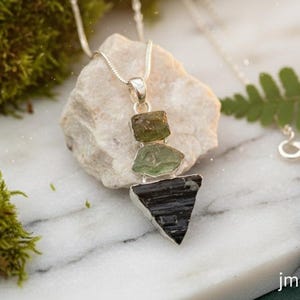 Puede incluir: Un collar de plata con un colgante de tres piedras en bruto apiladas: un peque&ntilde;o cristal verde oliva, un cristal verde claro m&aacute;s grande y una piedra negra triangular en la base. El colgante cuelga de una fina cadena de plata con cierre.