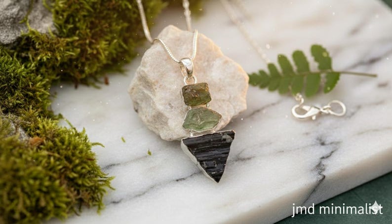 Puede incluir: Un collar de plata con un colgante de tres piedras en bruto apiladas: un peque&ntilde;o cristal verde oliva, un cristal verde claro m&aacute;s grande y una piedra negra triangular en la base. El colgante cuelga de una fina cadena de plata con cierre.