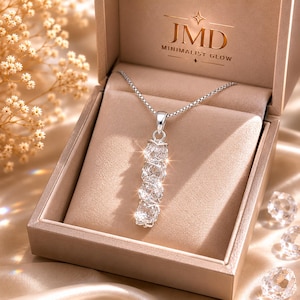 Puede incluir: Un collar de tono plateado con un colgante vertical que presenta cuatro piedras redondas, transparentes y facetadas dispuestas en columna. La joya se exhibe dentro de una caja beige con el texto JMD MINIMALIST GLOW en la tapa interior.