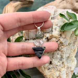 Puede incluir: Un collar con colgante de tres piedras apiladas suspendidas de una cadena de plata. La piedra superior es un peque&ntilde;o cristal rectangular verde oliva, la central es un cuarzo ovalado transparente y la inferior es una gran turmalina negra triangular.