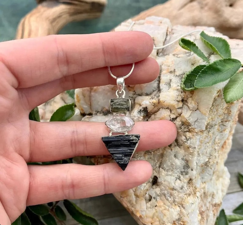 Puede incluir: Un collar con colgante de tres piedras apiladas suspendidas de una cadena de plata. La piedra superior es un peque&ntilde;o cristal rectangular verde oliva, la central es un cuarzo ovalado transparente y la inferior es una gran turmalina negra triangular.