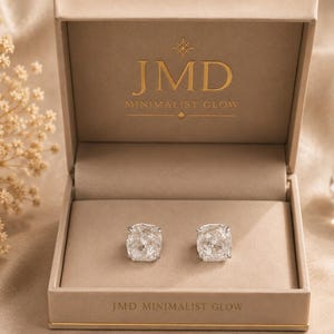 Puede incluir: Un par de pendientes de botón cuadrados con piedras transparentes facetadas engastadas en metal plateado, presentados dentro de un joyero rectangular beige. La tapa interior presenta el texto dorado JMD MINIMALIST GLOW con un icono de estrella.