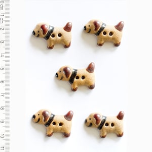 May include: Five ceramic dog buttons with brown and black details. The buttons are approximately 2 cm in size and have two holes for sewing.
