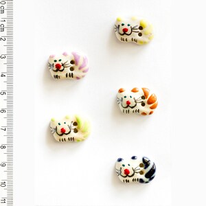 May include: Five ceramic cat buttons with different colored stripes. The buttons are white with painted details, including red noses, black whiskers, and green, blue, orange, yellow, and purple stripes.