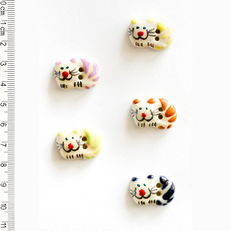 May include: Five ceramic cat buttons with different colored stripes. The buttons are white with painted details, including red noses, black whiskers, and green, blue, orange, yellow, and purple stripes.
