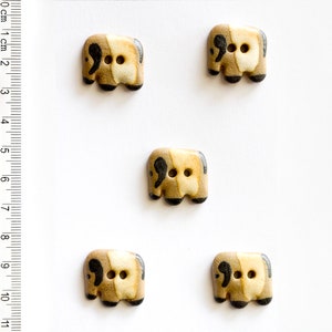 May include: Five ceramic buttons shaped like elephants. The buttons are brown and beige with black accents. Each button has two black eyes and a black trunk.