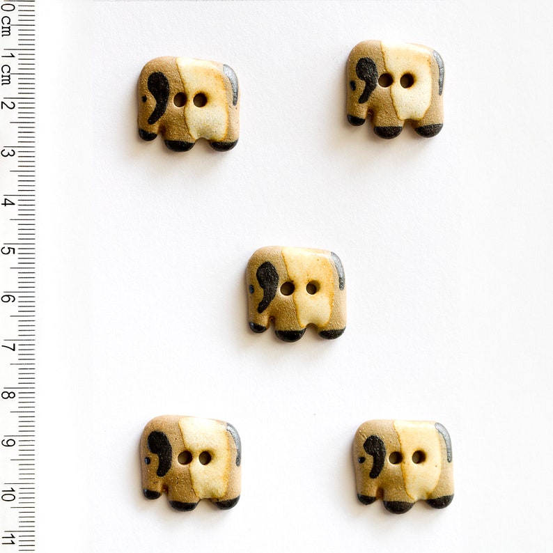 May include: Five ceramic buttons shaped like elephants. The buttons are brown and beige with black accents. Each button has two black eyes and a black trunk.