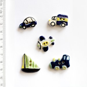 May include: Five ceramic buttons featuring different modes of transport. The buttons are blue, white and yellow with black outlines. The buttons are shaped like a car, a lorry, a plane, a sailboat and a train.
