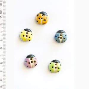 May include: Five ceramic ladybug buttons in different colours: yellow, blue, green, pink, and orange. Each button has black spots and a black head with two black dots for eyes.