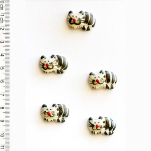 May include: Five ceramic buttons shaped like cats. The cats are white with black stripes and have red noses and black eyes. The buttons are about 1 cm in diameter.