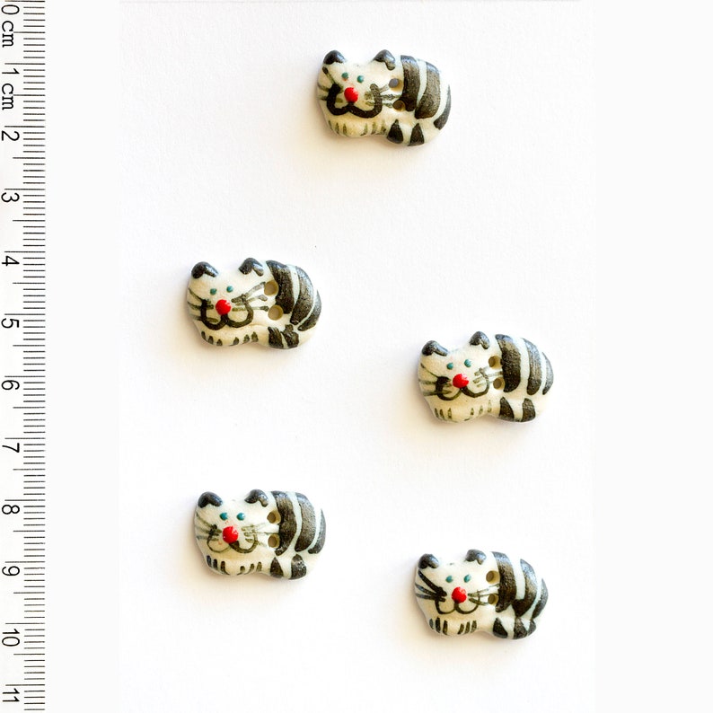 May include: Five ceramic buttons shaped like cats. The cats are white with black stripes and have red noses and black eyes. The buttons are about 1 cm in diameter.