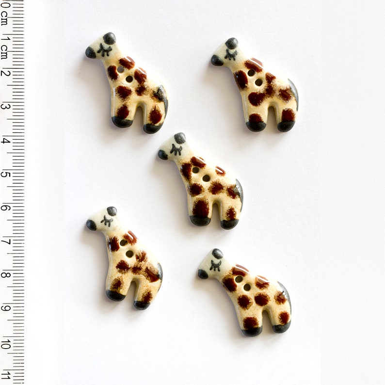 May include: Five ceramic buttons shaped like giraffes with brown spots on a white background. The buttons have black eyes and a black nose.