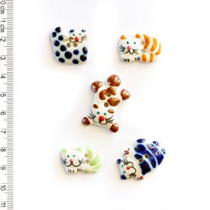 May include: Five ceramic cat buttons with different colored patterns. The buttons are white with brown, blue, orange, and green accents. The cats have black eyes and pink noses.
