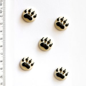 May include: Five white ceramic buttons with black bear paw prints. The buttons are approximately 1 inch in diameter.