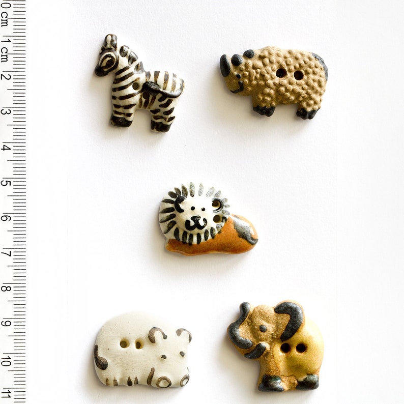May include: Five ceramic buttons featuring different animals: a zebra, a rhino, a lion, a hippopotamus, and a bear. The buttons are all white with black and brown details. They are approximately 1 inch in diameter.