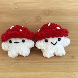May include: Two crocheted mushroom plushies with white bodies and red and white spotted caps. The mushrooms have black eyes and smiling mouths.