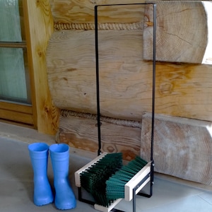 Pode incluir: Um par de botas de borracha azul claro ao lado de um limpador de botas de metal preto com cerdas verdes. O limpador de botas tem uma estrutura de madeira. O fundo apresenta uma parede de madeira.