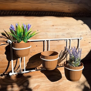 Op de afbeelding: Drie bruine, geribbelde bloempotten met witte rand hangen aan een wit metalen rek. De potten bevatten kunstplanten met paarse bloemen en groene bladeren. Het rek is op een houten muur gemonteerd.