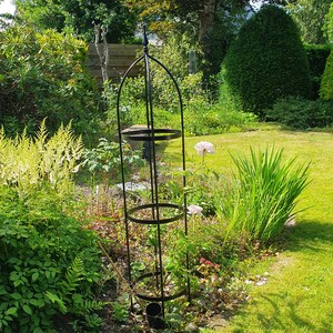 Op de afbeelding: Een hoge, zwarte metalen obelisk-plantensteun met een puntige knop aan de bovenkant en drie cirkelvormige steunringen verbonden door verticale staven. De structuur is ontworpen om klimplanten te ondersteunen en staat rechtop in de grond.