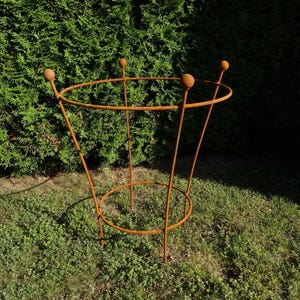 Puede incluir: Un soporte para plantas de metal color óxido con un anillo superior circular y una base circular más pequeña conectados por cuatro varillas verticales. Cada varilla se extiende hacia arriba más allá del anillo superior y termina en una pequeña esfera.