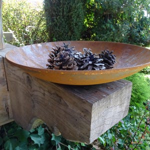 Op de afbeelding: Een roestige metalen kom met een brede rand zit op een verweerde houten balk. De kom is gevuld met dennenappels, waarvan sommige bruin en andere grijs zijn.