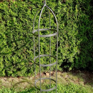 Op de afbeelding: Een hoge, cilindervormige tuinobelisk van grijs metaal, voorzien van een puntige top, drie horizontale cirkelvormige ringen en verticale steunstaven die in de grond steken om een stabiele structuur te bieden voor klimplanten.