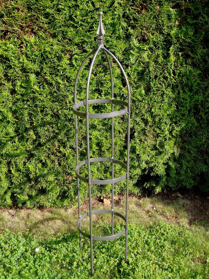 Op de afbeelding: Een hoge, cilindervormige tuinobelisk van grijs metaal, voorzien van een puntige top, drie horizontale cirkelvormige ringen en verticale steunstaven die in de grond steken om een stabiele structuur te bieden voor klimplanten.