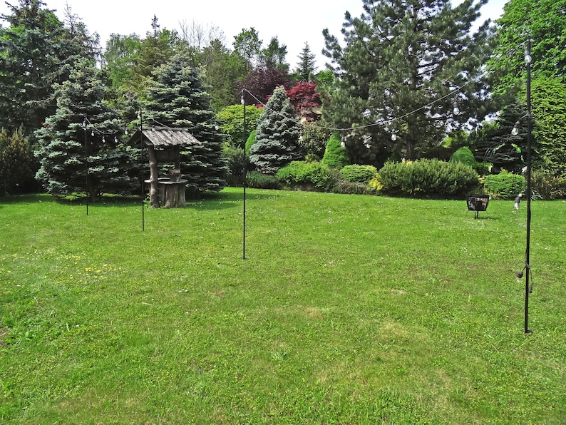 Puede incluir: Una escena de jard&iacute;n con un pozo de madera, hierba verde y varios postes de metal negro con luces de cadena tendidas entre ellos.