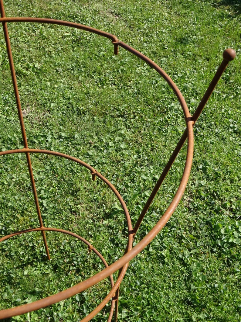 Op de afbeelding: Een roestige metalen plantenondersteuning met drie niveaus. De ondersteuning is gemaakt van ronde metalen staven en heeft een afgeronde bovenkant.