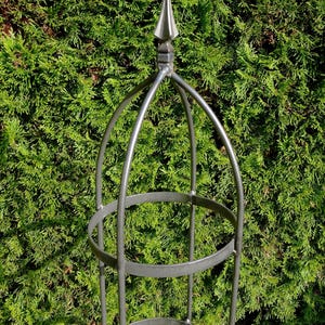 Op de afbeelding: Een hoge, cilindervormige tuinobelisk van donkergrijs metaal. De structuur bestaat uit verticale staven verbonden door twee horizontale cirkelvormige ringen, bekroond met een puntige finial. Het frame is open voor klimmende planten.