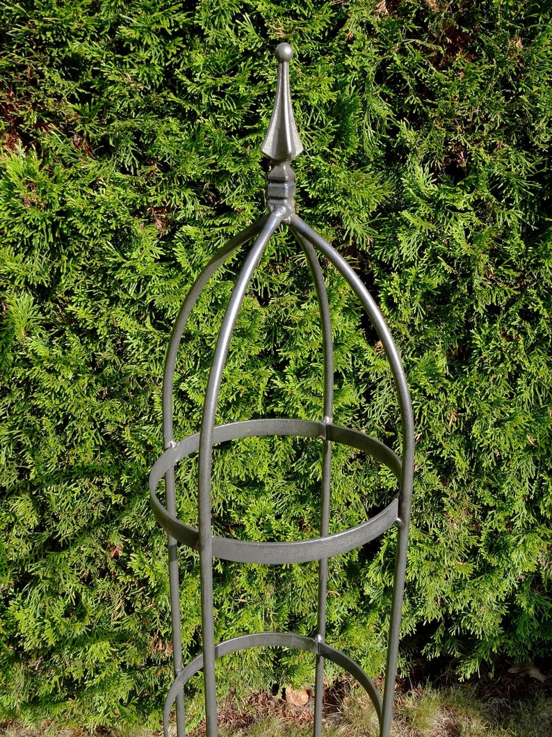 Op de afbeelding: Een hoge, cilindervormige tuinobelisk van donkergrijs metaal. De structuur bestaat uit verticale staven verbonden door twee horizontale cirkelvormige ringen, bekroond met een puntige finial. Het frame is open voor klimmende planten.