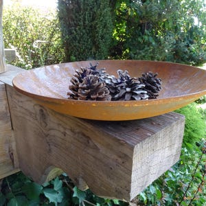 Op de afbeelding: Een roestige metalen kom gevuld met dennenappels staat op een houten balk. De kom is ovaalvormig en heeft een licht gerimpelde oppervlak. De dennenappels zijn bruin en hebben een natuurlijke, rustieke uitstraling.