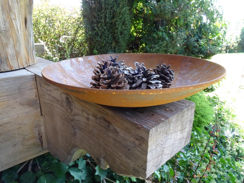 Op de afbeelding: Een roestige metalen kom gevuld met dennenappels staat op een houten balk. De kom is ovaalvormig en heeft een licht gerimpelde oppervlak. De dennenappels zijn bruin en hebben een natuurlijke, rustieke uitstraling.