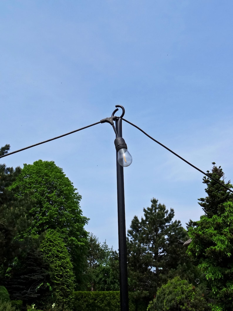 Puede incluir: Un poste de luz de metal negro con una sola bombilla colgando de &eacute;l. El poste est&aacute; en un &aacute;rea cubierta de hierba con &aacute;rboles en el fondo.