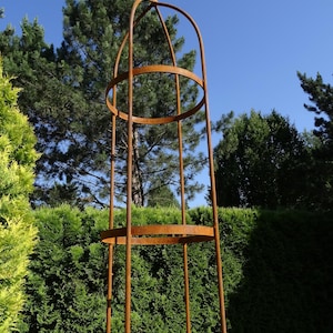 Op de afbeelding: Een hoge, cilindervormige tuinobelisk van roestig metaal. De structuur bestaat uit verticale staven verbonden door twee horizontale cirkelvormige ringen, bekroond met een puntige finial. Het ontwerp is open en kooiachtig voor klimplanten.