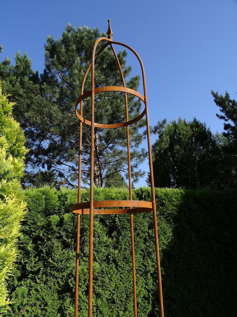 Op de afbeelding: Een hoge, cilindervormige tuinobelisk van roestig metaal. De structuur bestaat uit verticale staven verbonden door twee horizontale cirkelvormige ringen, bekroond met een puntige finial. Het ontwerp is open en kooiachtig voor klimplanten.