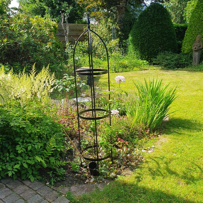 Op de afbeelding: Een hoge, zwarte metalen obelisk-plantensteun met een puntige knop aan de bovenkant en drie cirkelvormige steunringen verbonden door verticale staven. De structuur is ontworpen om klimplanten te ondersteunen en staat rechtop in de grond.