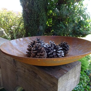 Op de afbeelding: Een roestige metalen kom met een brede rand staat op een houten oppervlak. De kom is gevuld met vier dennenappels.