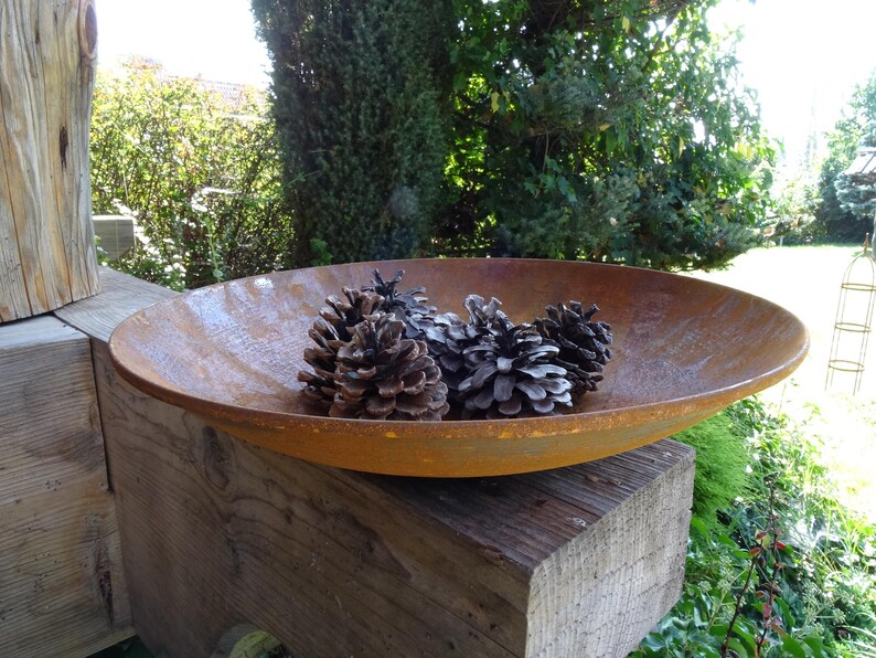 Op de afbeelding: Een roestige metalen kom met een brede rand staat op een houten oppervlak. De kom is gevuld met vier dennenappels.